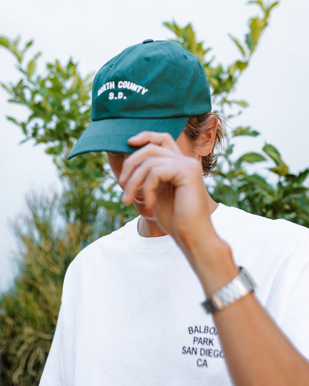 Person wearing a green cap with 'North County S.D.' text and a white t-shirt with 'Balboa Park San Diego CA' text, outdoors.