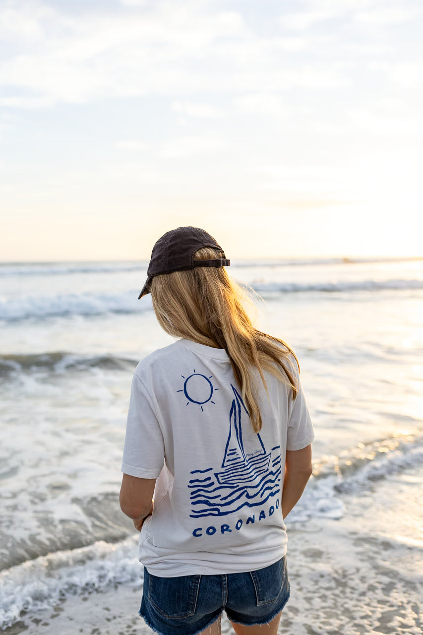 May Gray model wearing a white t-shirt with a sailboat design and 'Coronado' text, standing on a beach at Oceanside Harbor